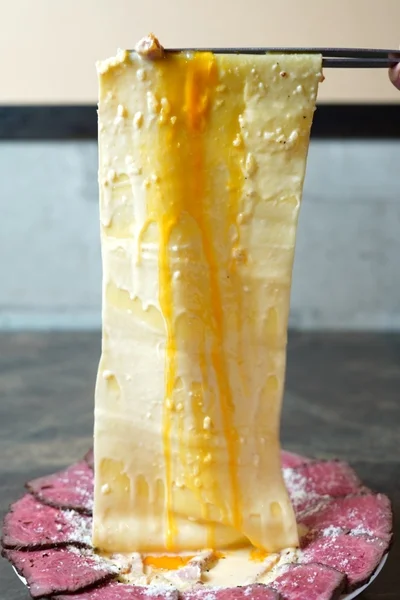 箸で持ち上げられたとろけるチーズのような塊から卵黄が滴り落ち、下には薄切りのローストビーフが敷き詰められた、食欲をそそる創作料理のクローズアップ。