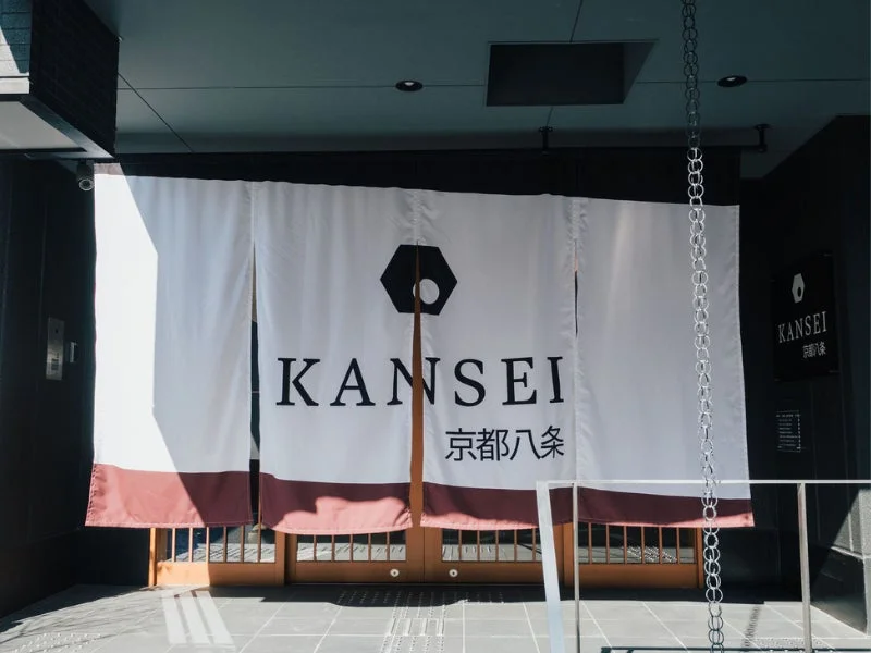 京都八条にある「KANSEI」のエントランス。白い暖簾には施設名とロゴが大きく印字され、モダンで洗練された和の雰囲気を醸し出している。