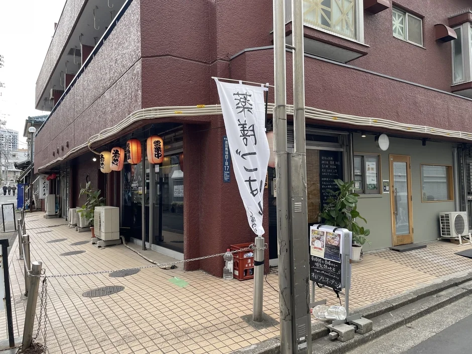日本の街角にある、茶色い建物の一階部分に位置する「喫茶朝ごこち」というカフェまたは飲食店を捉えた写真です。入口には赤い提灯や白い旗が飾られ、営業中であることが示唆されています。入り口付近の黒板には「本日プレオープン」と書かれており、開店したばかりの様子がうかがえます。