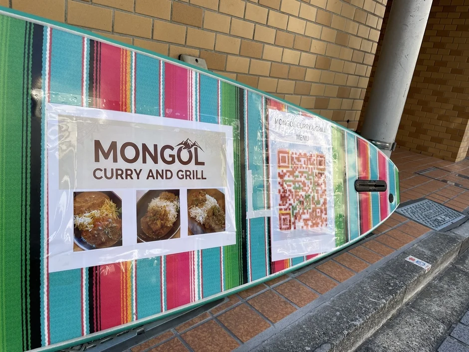 カラフルなストライプ模様のボードに「MONGOL CURRY AND GRILL」の文字と料理の写真、QRコードが貼られた看板。