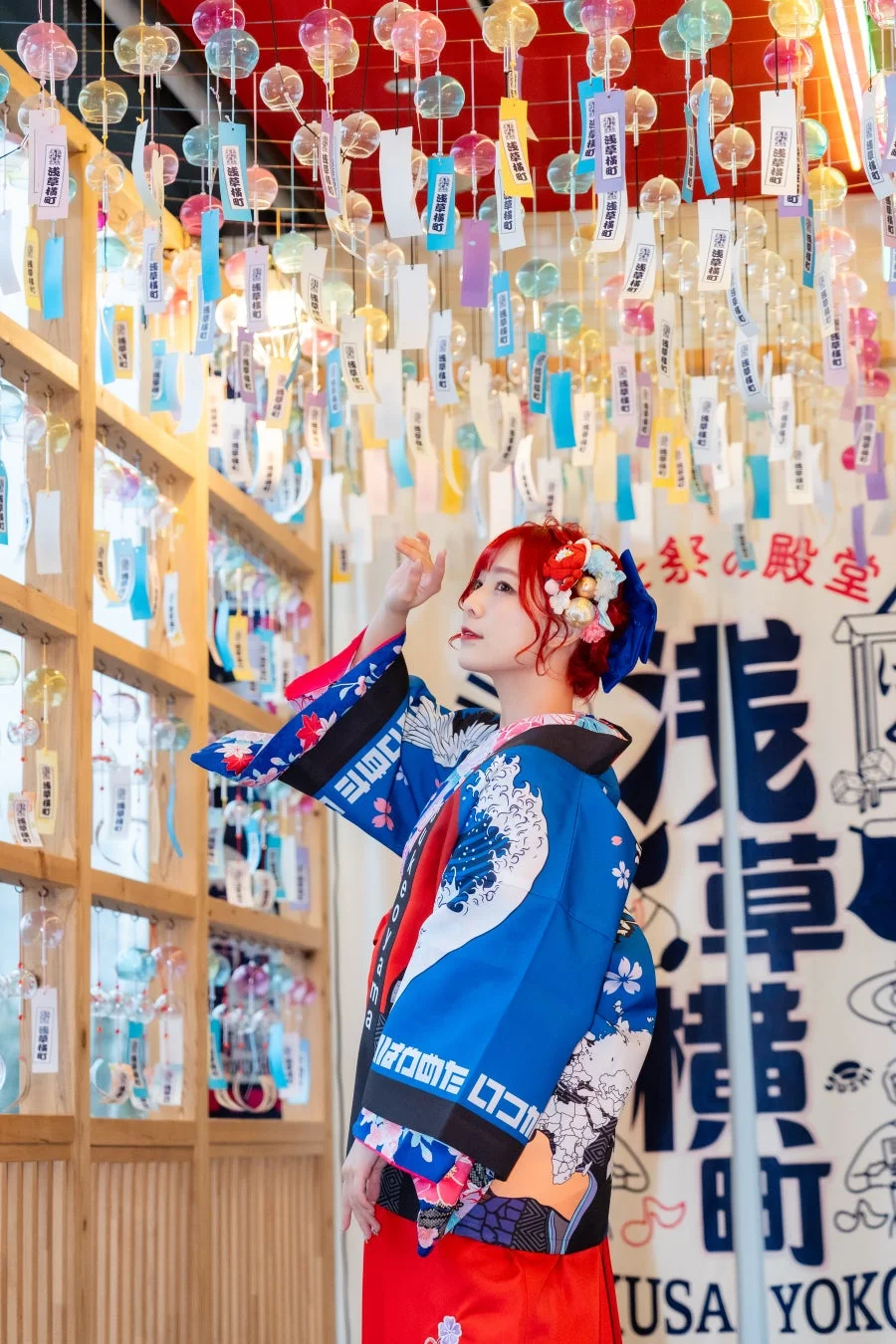 カラフルな風鈴が飾られた空間で、鮮やかな着物を着た赤髪の女性が上を見上げている