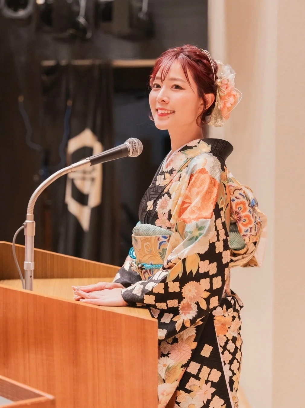 華やかな着物を着た女性が演台の前に立ち、マイクに向かって笑顔で話している様子