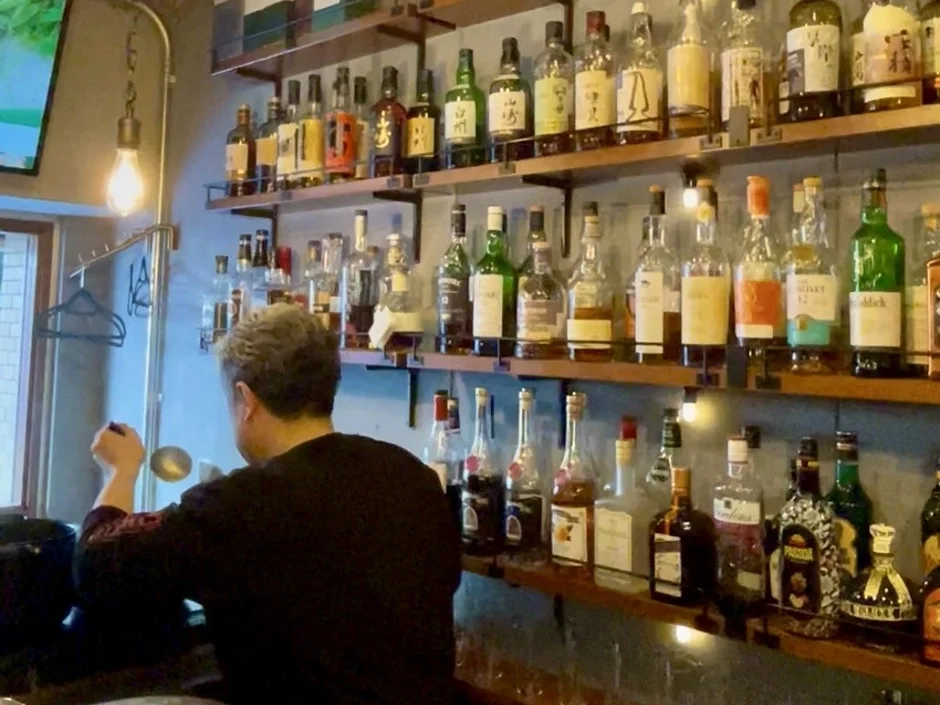 バーカウンターで背中を向けた人物が作業しており、その背後には様々な種類のウイスキーや洋酒のボトルがずらりと並んだ棚が印象的です。