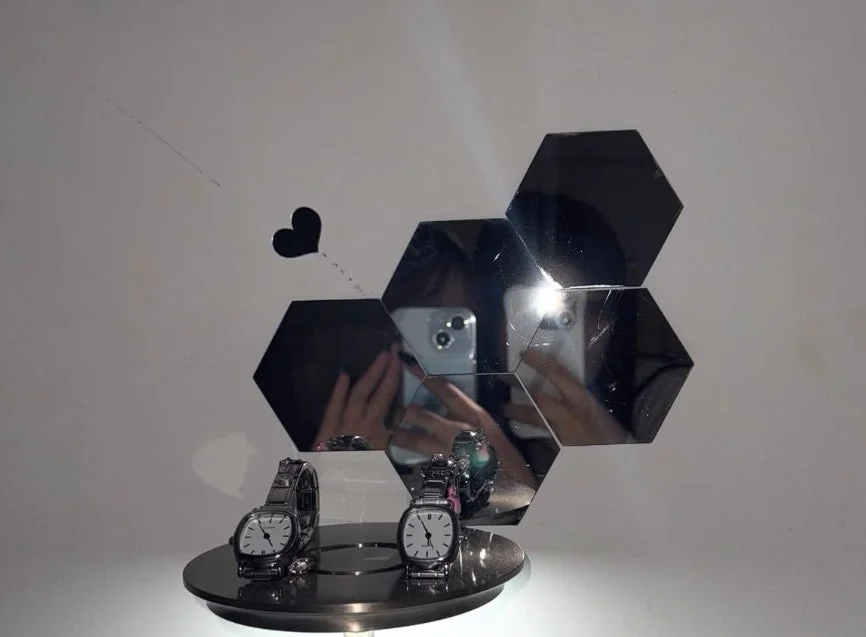 白い壁を背景に、ディスプレイスタンドに置かれた2つの腕時計が写っています。壁には六角形のミラーが複数配置され、撮影者のスマートフォンと手が映り込んでいます。小さなハートの飾りも添えられています。