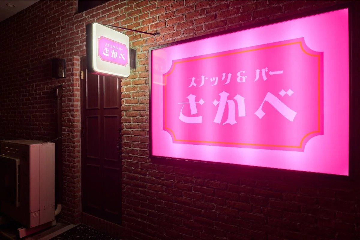 レンガの壁に設置された「スナック&バー さかべ」のネオンサインが鮮やかに輝く夜の店舗外観。レトロな雰囲気のバーの入口が、温かい光で照らされています。