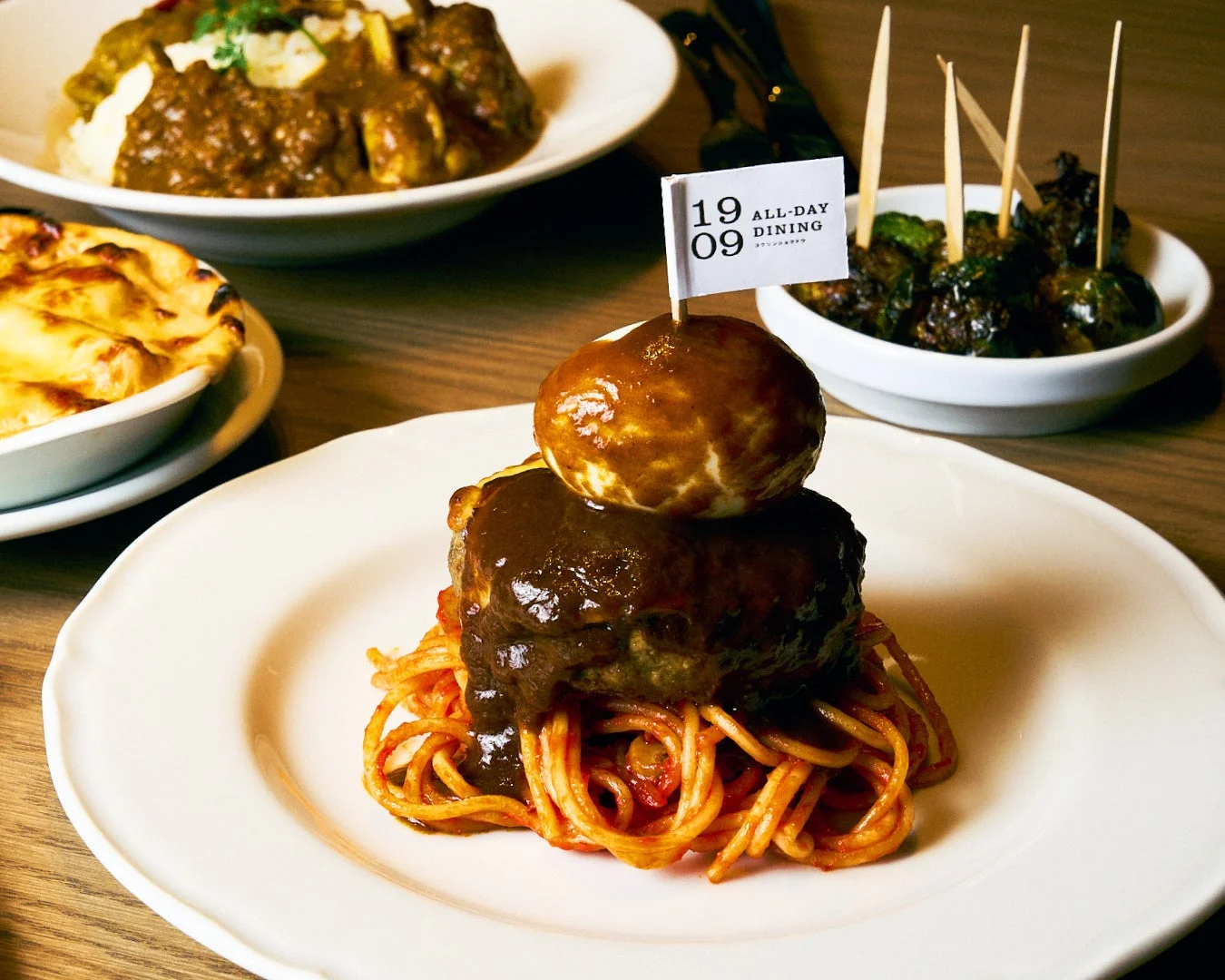 ハンバーグと半熟卵が乗ったスパゲッティをメインに、カレーやグラタンなどの洋食が並ぶ食卓の様子。旗には店名らしき「19 09 ALL-DAY DINING スプリングテラス」の文字が見える。