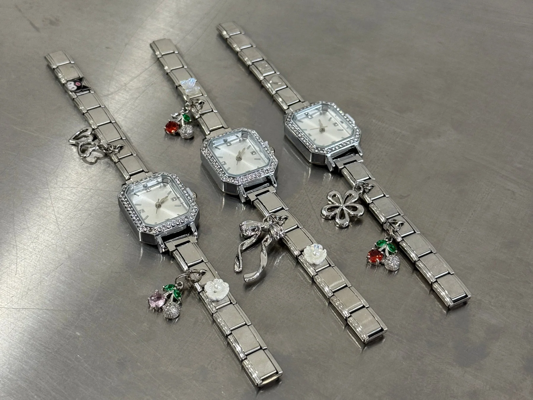 シルバーのブレスレットウォッチが3本並べられています。長方形の文字盤にはラインストーンが施され、バンドにはチェリー、リボン、花、ハートなどの可愛らしいチャームが付いています。