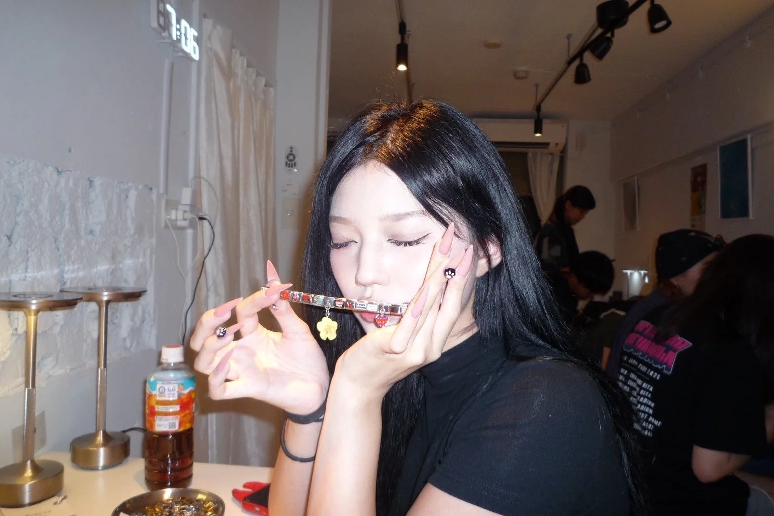 長い黒髪の若い女性が、長いネイルを施した指でチャームブレスレットを口元に持っていく様子を捉えた画像です。カフェのような屋内の空間で、他の人々も写り込んでいます。