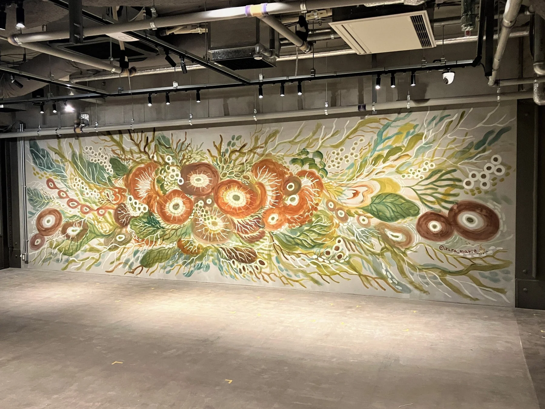 露出した配管や照明のあるモダンな空間に、オレンジや緑を基調とした有機的な花や植物を思わせる大規模な壁画が描かれています。右下には「@ikura_marie」のサインがあります。
