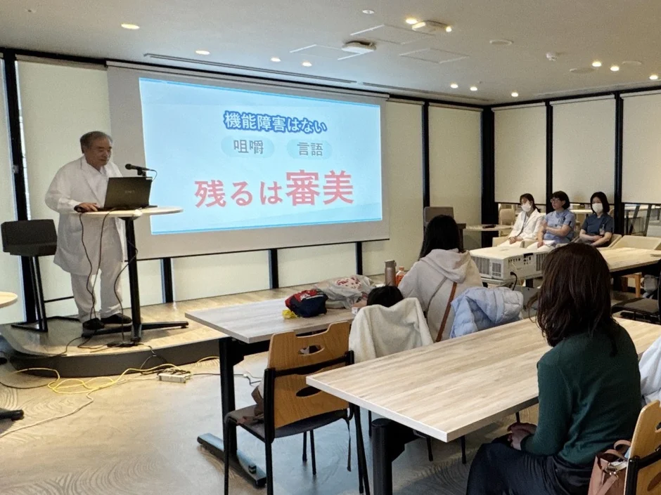 白衣を着た講師がプロジェクターを使って聴衆にプレゼンテーションを行っている様子です。スクリーンには「機能障害はない」「咀嚼」「言語」「残るは審美」と表示されており、医療や歯科関連の専門的な内容の講演会と推測されます。