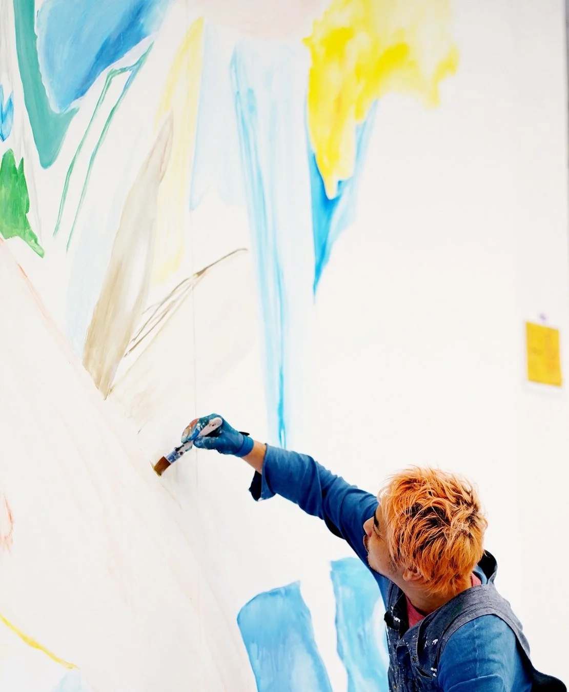 オレンジ色の髪の人物が、青い手袋をはめて大きな白い壁にカラフルな抽象画を描いている様子。集中して筆を動かし、鮮やかな色彩が広がるダイナミックな創作活動の一場面です。