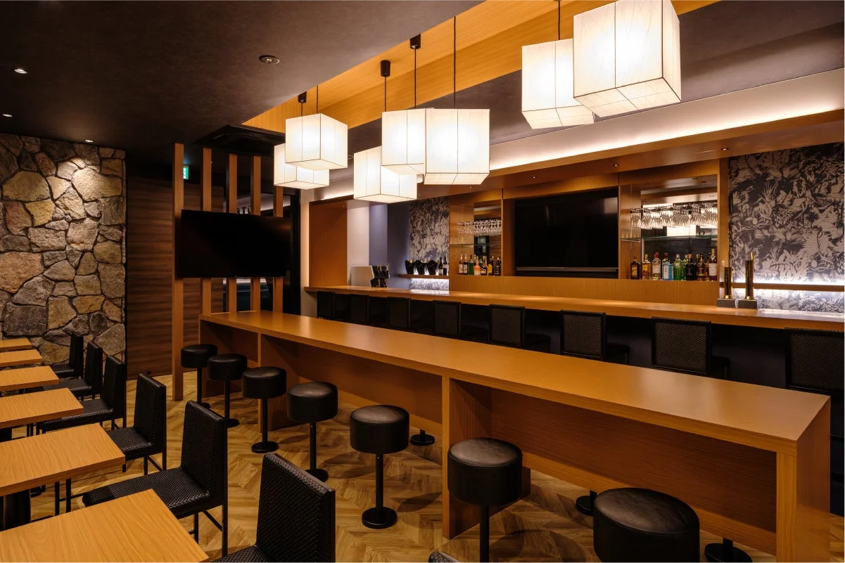 現代的な和風のバーまたはラウンジの内部。長い木製のカウンターと黒いバースツールが並び、天井からは和紙のようなペンダントライトが吊るされている。石壁のアクセントや温かみのある照明が、落ち着いた洗練された空間を演出している。
