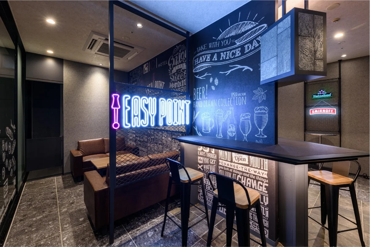 ネオンサインが光るモダンなバーラウンジ。チョークアートで飾られた壁とカウンター、そしてソファ席があり、リラックスできる空間を提供しています。ビールやカクテルを楽しむのに最適な雰囲気です。