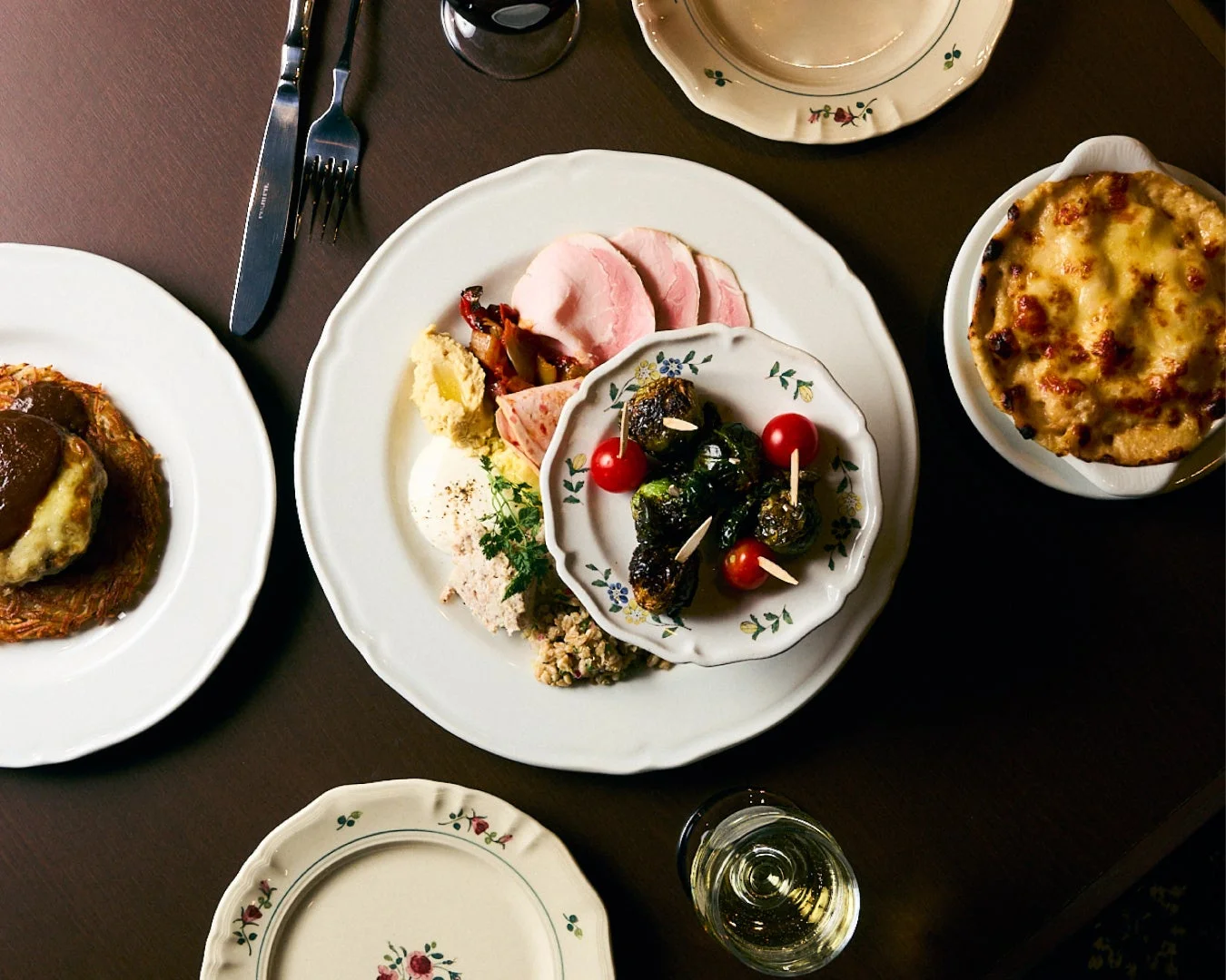 暗い木製のテーブルに、ハムの盛り合わせ、ハンバーグ、グラタンなど、様々な種類の洋食が並べられています。白ワインのグラスも添えられ、豪華なディナーの様子が伺えます。