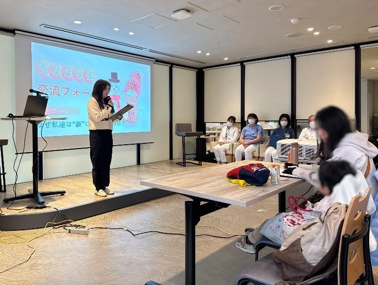 室内で開催されている交流フォーラムで、一人の女性がマイクを持ってプロジェクターに映し出された資料を使いながら発表しています。複数の参加者が熱心に耳を傾けています。