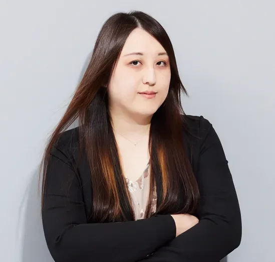 グレーの背景を背に、黒いブレザーと柄物のトップスを着用した長髪の女性が腕を組み、カメラを見つめているポートレート写真。真剣な表情を浮かべている。