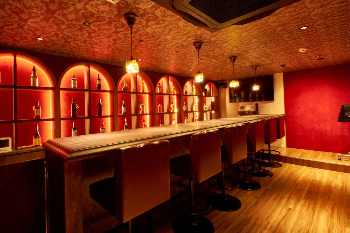 赤と金色の豪華な内装が特徴的なバーの店内です。長いカウンターとバーチェアが並び、奥にはボトルが多数飾られた間接照明付きの棚があります。落ち着いた雰囲気で、大人の空間を演出しています。