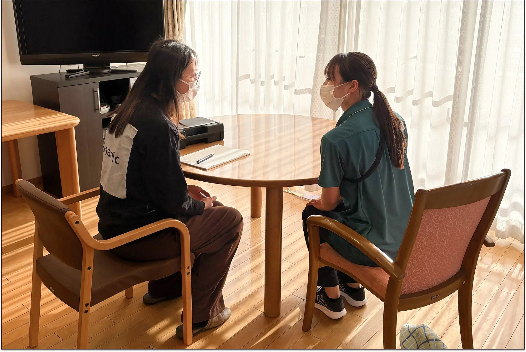 マスクを着用した2人の女性が丸いテーブルを挟んで向かい合い、会話している様子。室内にはテレビやカーテンがあり、落ち着いた雰囲気の中で何らかの打ち合わせや相談が行われているように見えます。