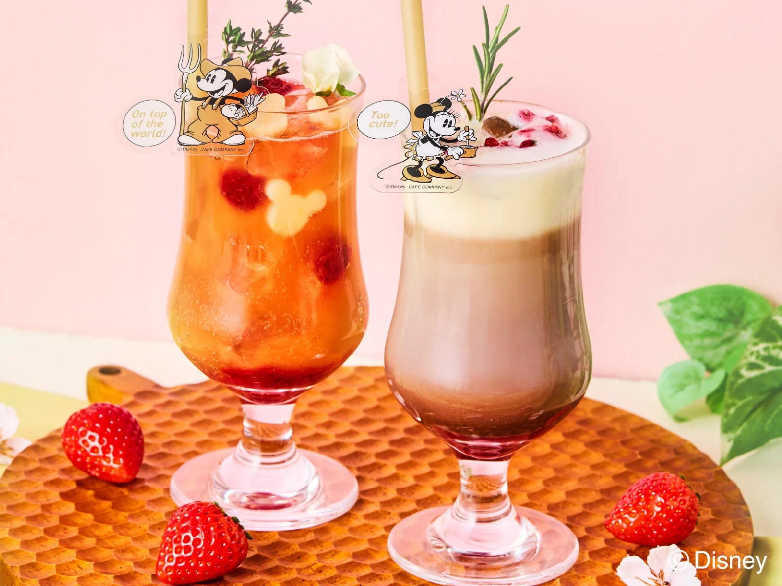 ピンクの背景に木製トレイが置かれ、ミッキーマウスとミニーマウスの飾りがついた2種類のカラフルなデザートドリンクが並んでいます。周りには新鮮なイチゴが添えられており、可愛らしいカフェメニューの様子を捉えた画像です。