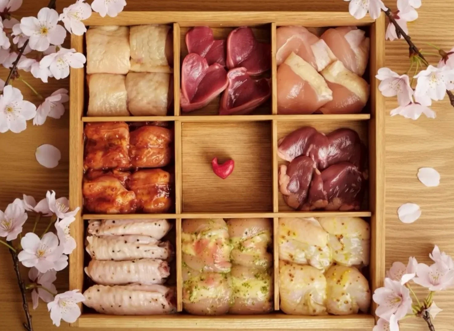 木製の箱に鶏肉の様々な部位と内臓が美しく盛り付けられています。一部は味付けされており、中央には赤いハート、周りには桜の花が添えられています。春の訪れを感じさせる、贈り物やお祝いに最適な華やかな肉の詰め合わせです。