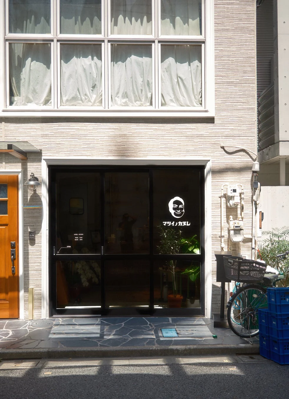 日本の都市部にある店舗の正面。暗いガラス扉には「マツイノカズレ」という店名と顔のロゴ、そして営業時間が白く表示されている。建物の外壁は明るい色で、上階には白いカーテンがかかった窓があり、店の前には自転車が停められている。