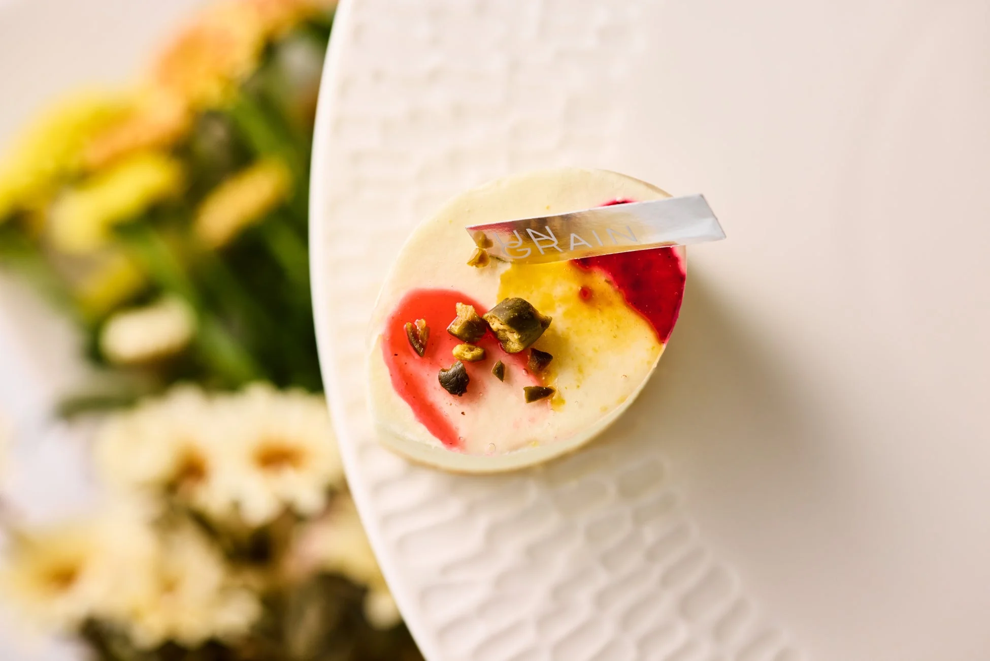 白い皿に盛られた、赤と黄色の模様が美しい丸いデザート。刻んだピスタチオがトッピングされ、背景にはぼかした黄色い花が見える、上品な一品です。