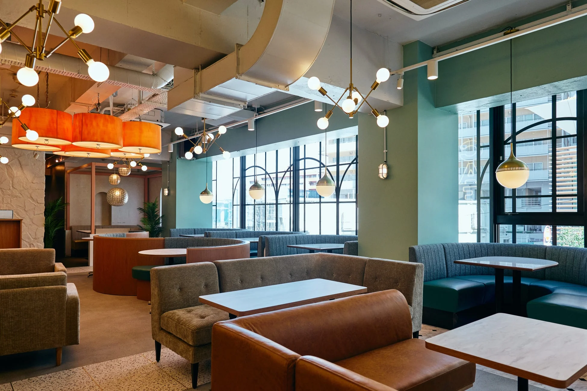 モダンでおしゃれなカフェまたはレストランの店内風景。様々なタイプのソファ席やブース席が配置され、ユニークなデザインの照明器具が空間を彩っています。窓からは都市の景色が見え、落ち着いた雰囲気でくつろげる空間が広がっています。