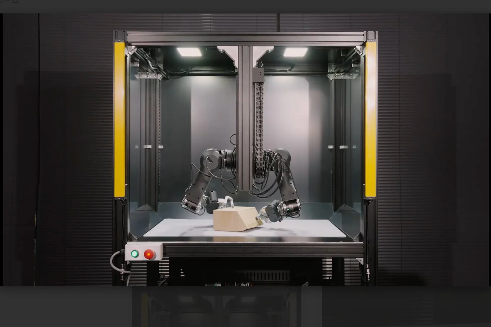 産業用のワークセル内で、2本のロボットアームが台上の段ボール箱を操作している様子を捉えた画像です。