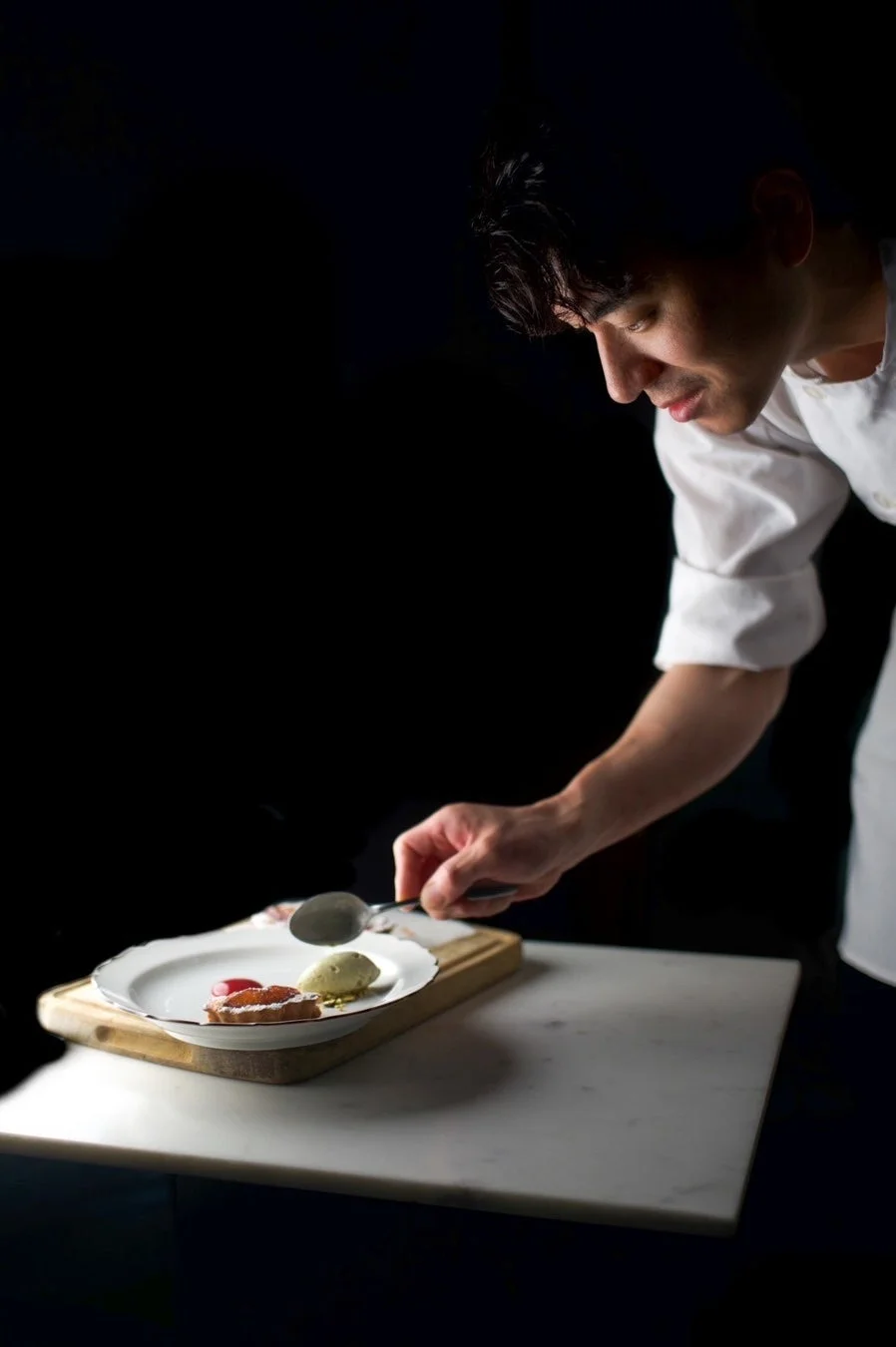 暗い背景の中、スポットライトを浴びたシェフが集中してデザートを盛り付けている様子。