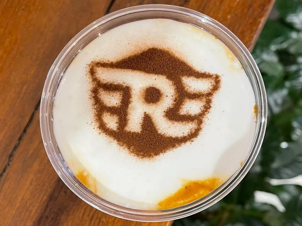 ミルクフォームの上にココアパウダーで「R」の文字と翼のようなデザインのラテアート