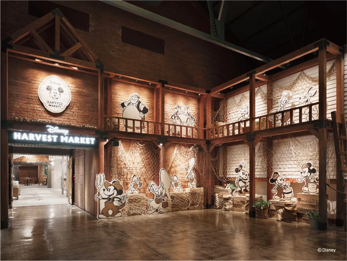 ディズニーハーベストマーケットのエントランスと店内ディスプレイ。木造とレンガの壁が素朴な雰囲気を醸し出し、ミッキーマウスなどのディズニーキャラクターが収穫や漁をテーマにした衣装で飾られています。漁網や藁が使われた暖かく魅力的な空間です。