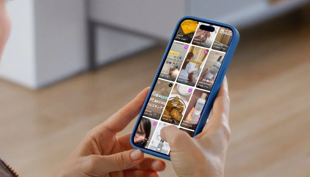 青いケースのスマートフォンを操作している人物が写っています。画面には、美容製品や食べ物、ライフスタイルに関する画像がグリッド表示されており、日本語のテキストや数字が見えます。