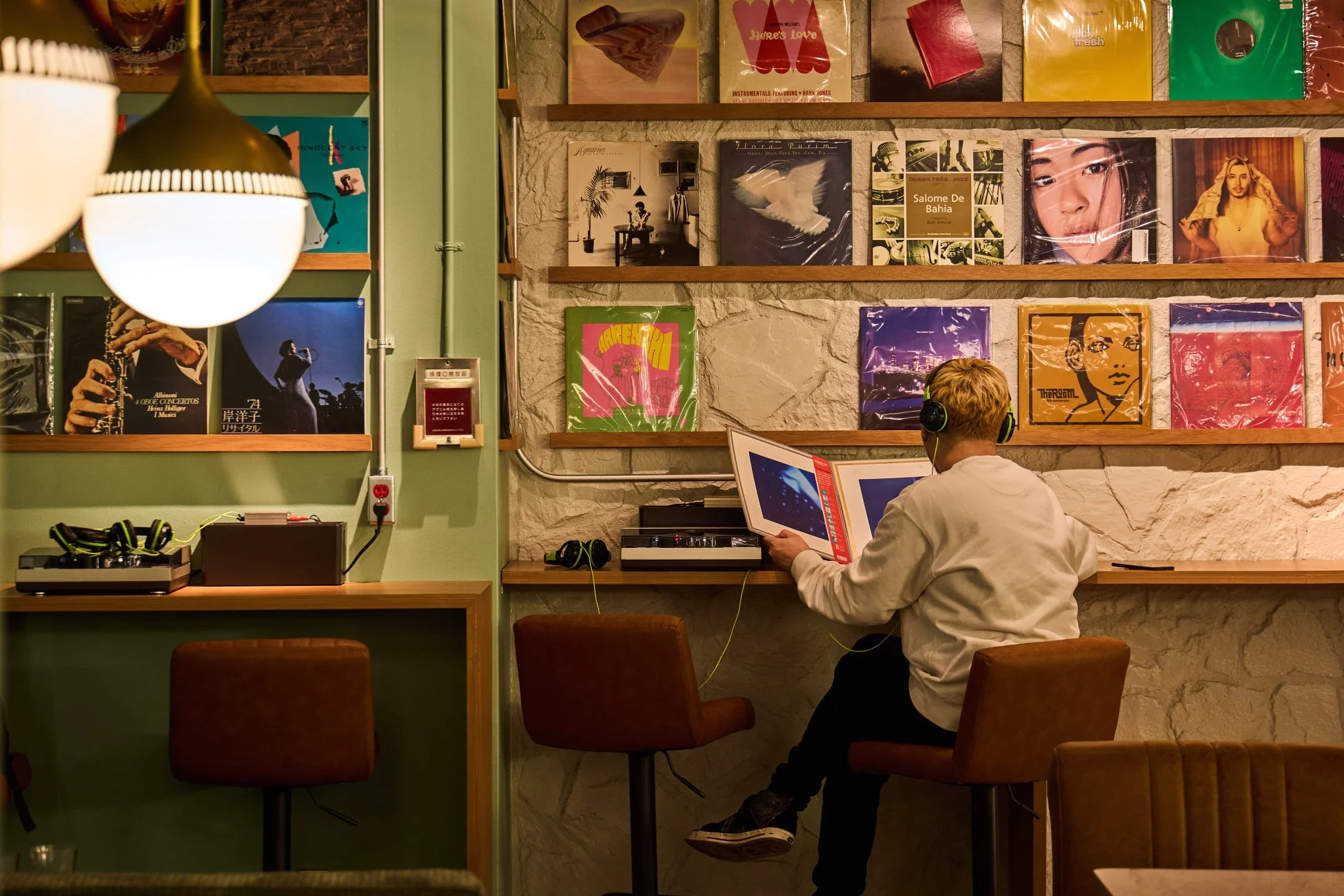 レコードがずらりと並んだ空間で、ヘッドホンをつけた男性がレコードを手に音楽を楽しんでいる。暖色系の照明が心地よい、落ち着いた雰囲気のリスニングバーの様子。