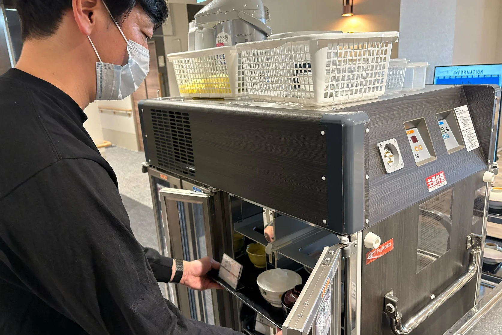 マスクを着用した男性が、配膳用の保温カートから食器を取り出している様子です。カートの上には炊飯器やカゴが置かれ、施設での食事提供の準備をしている場面を捉えています。