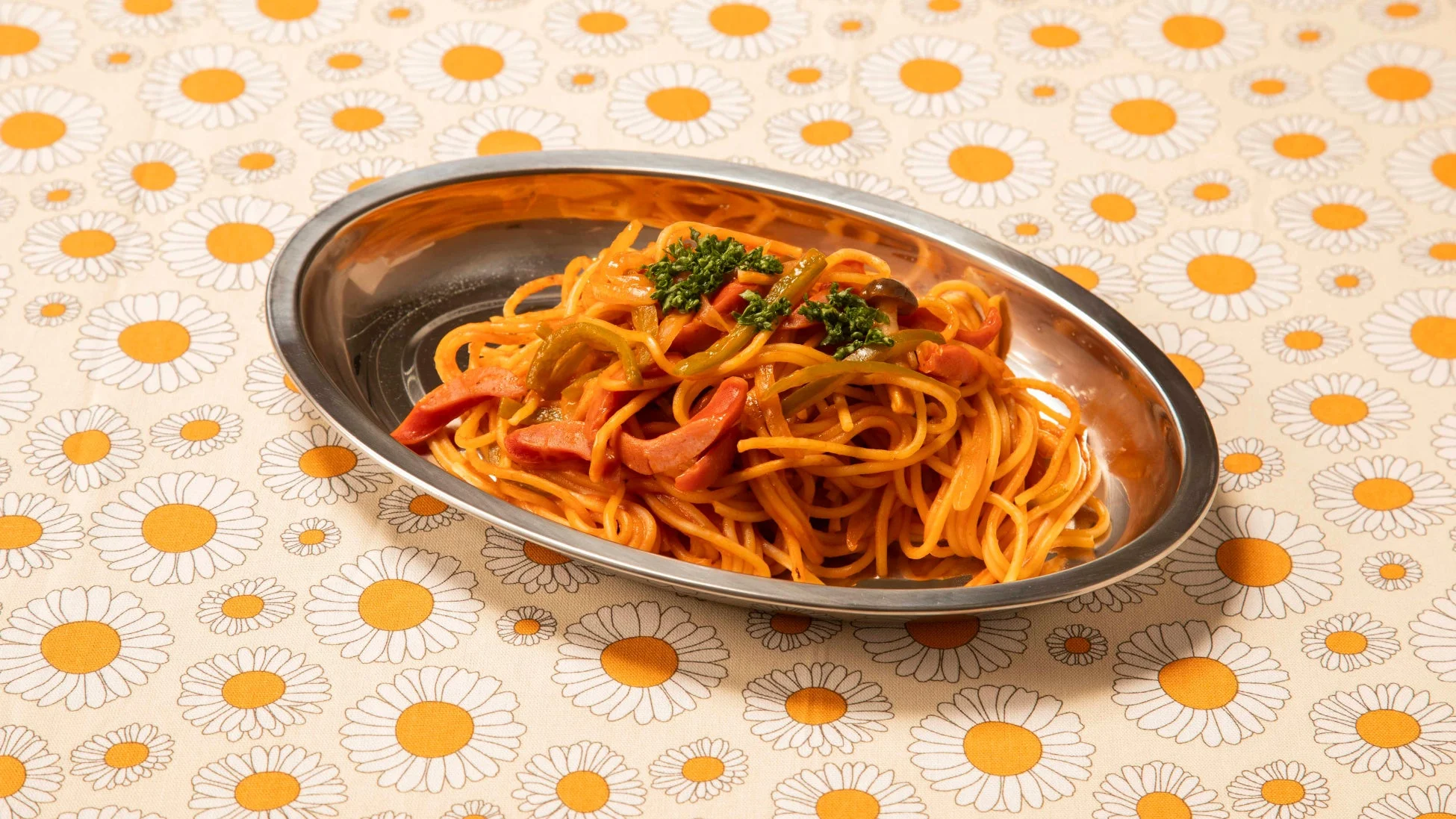 デイジー柄のテーブルクロスの上に、銀色の楕円皿に盛られた昔ながらのナポリタン。ソーセージやピーマンが彩り豊かに添えられ、レトロな喫茶店の定番メニューを思わせる一品です。