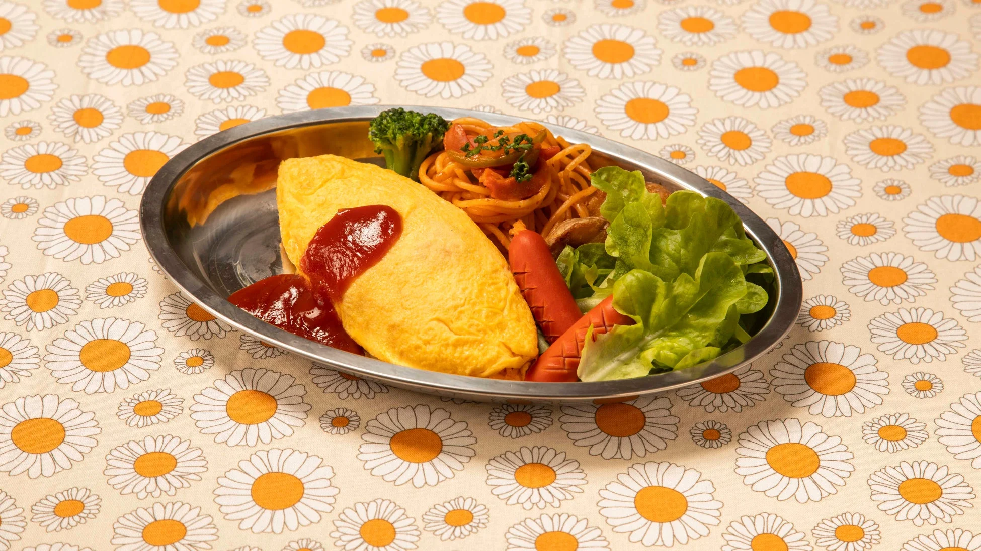 ケチャップがかかったオムライスを中心に、ナポリタン、ウインナー、サラダ、ブロッコリーが盛り付けられた洋食プレート。デイジー柄のレトロなテーブルクロスの上で、懐かしい雰囲気の食事が提供されています。