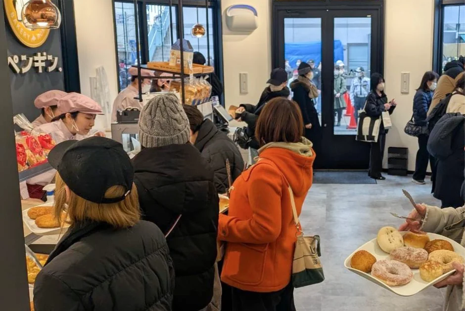 賑わうパン屋の店内で、多くの客がパンやドーナツを選び、レジに並んでいます