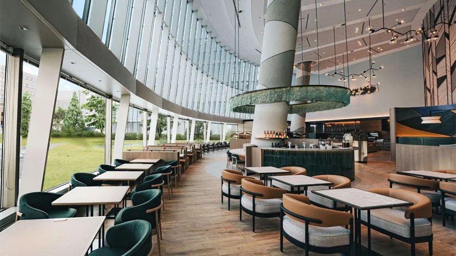 大きな窓から緑が見える、広々としたモダンなカフェまたはレストランの内部。様々なデザインの座席が配置され、開放的で洗練された雰囲気です。