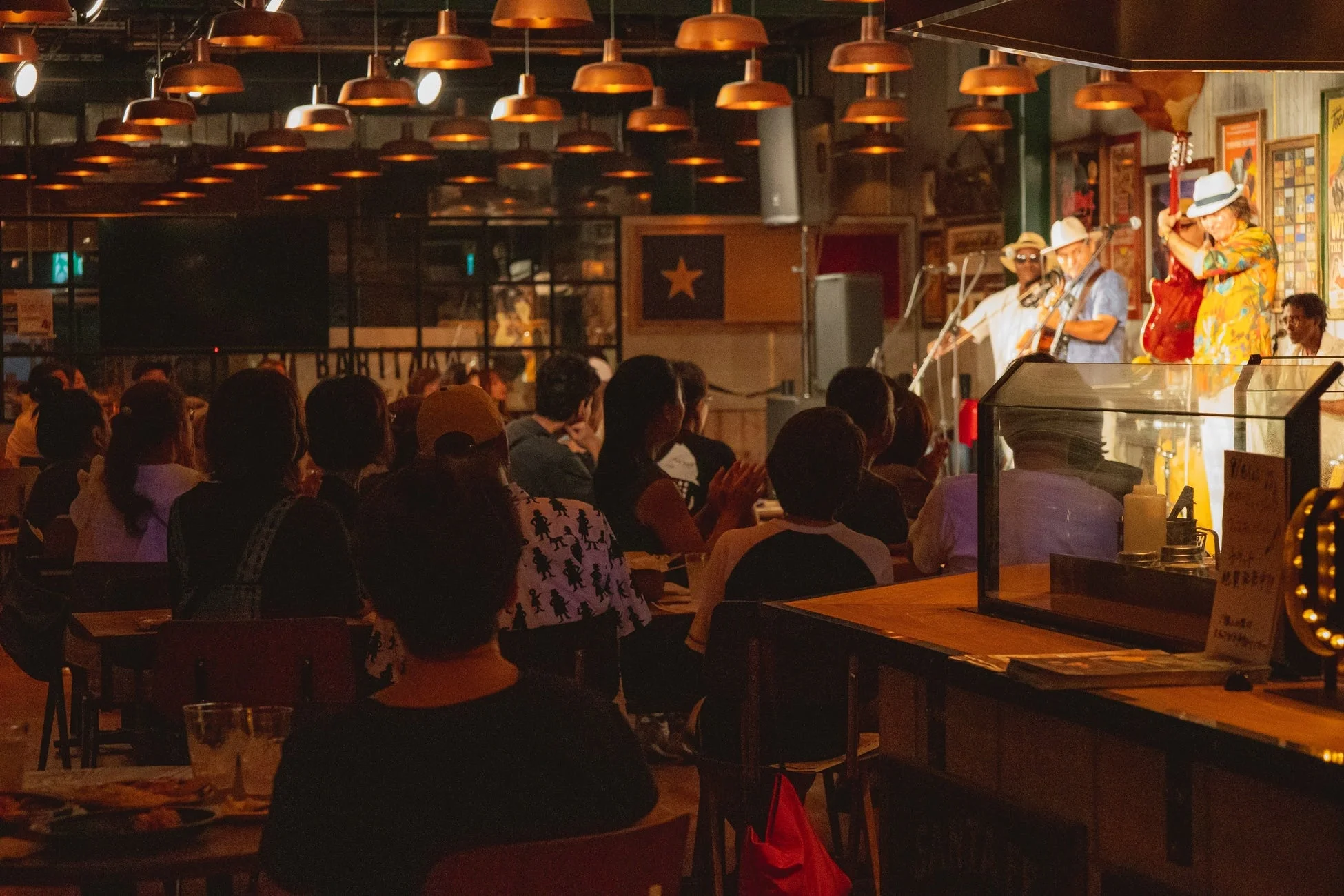 温かい照明のバーやレストランで、バンドがライブ演奏を行っており、多くの観客がその音楽を楽しんでいる夜のシーン。