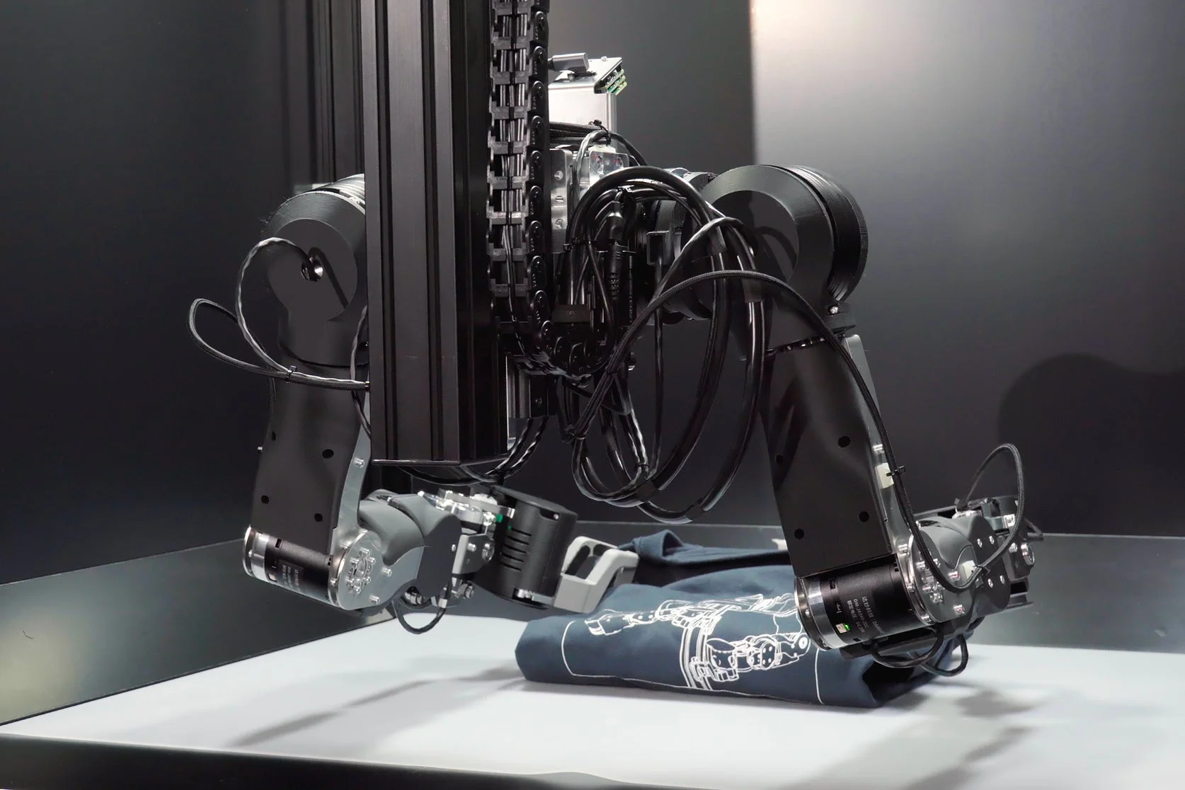 精密なロボットアームが、ロボットのイラストが描かれた青いTシャツと接している様子を捉えた画像です。自動化された作業風景や、産業用ロボットの導入事例を示唆しています。