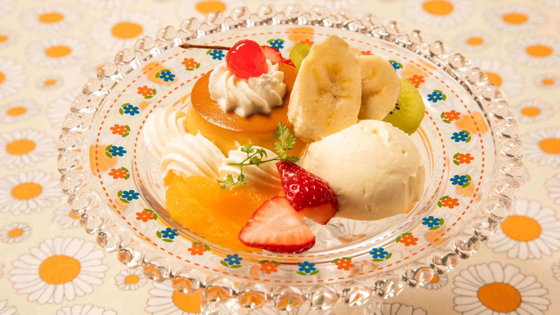 花柄のテーブルクロスに置かれたガラス皿のプリンアラモード。プリン、ホイップクリーム、バナナ、いちご、キウイ、オレンジ、アイスクリームが彩り豊かに盛り付けられ、チェリーがアクセントになった可愛らしいデザートです。