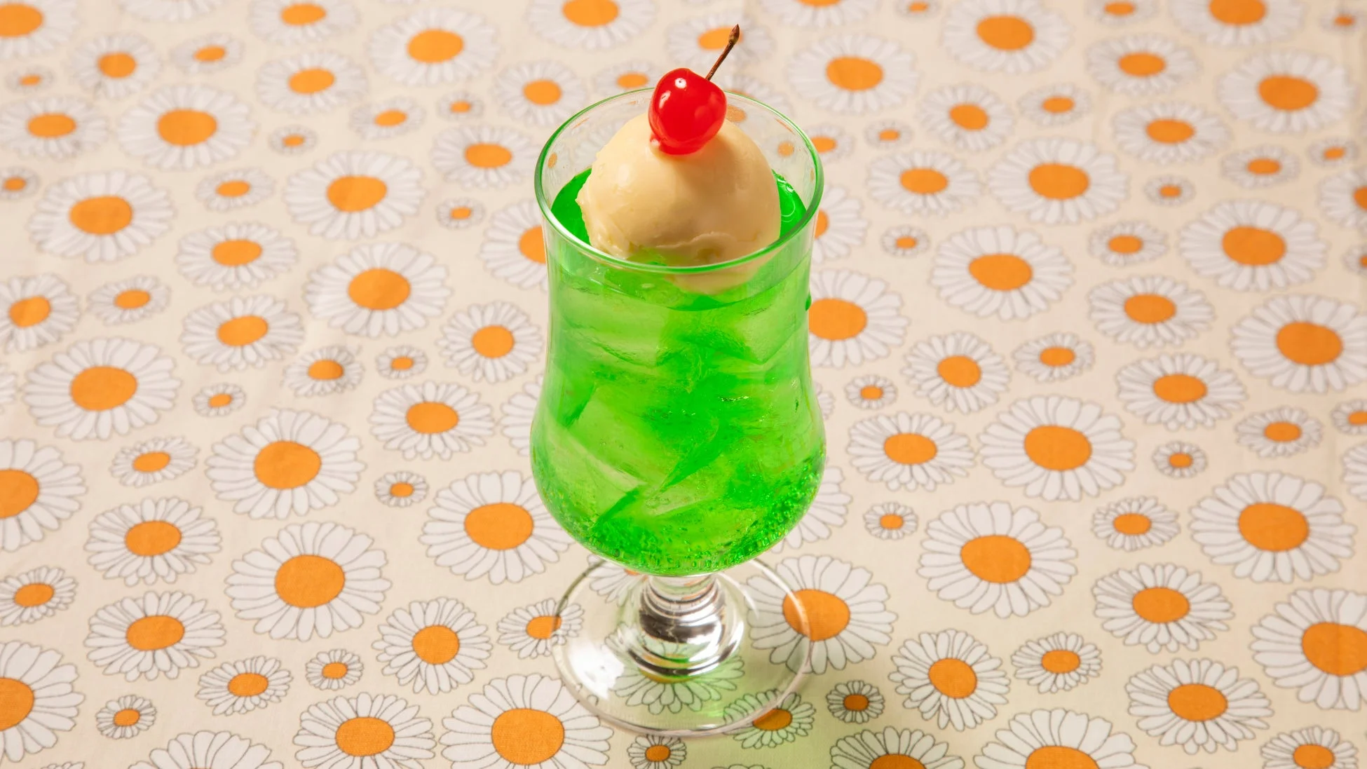 緑色のメロンソーダにバニラアイスと真っ赤なチェリーが乗ったクリームソーダです。デイジー柄のテーブルクロスに置かれ、レトロで涼しげな雰囲気を感じさせます。