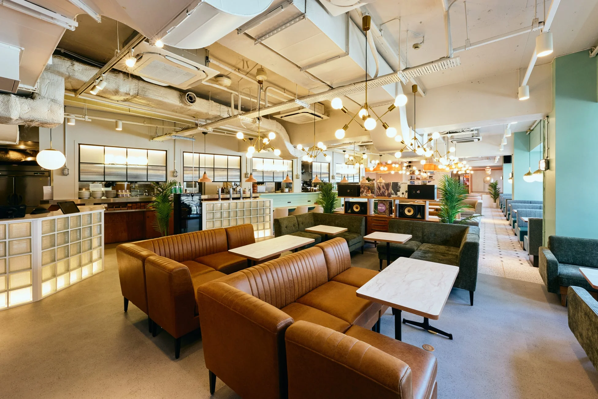 モダンで広々としたカフェまたはラウンジの室内。茶色と緑のソファ席、大理石調テーブルが並び、天井の配管と特徴的な照明がスタイリッシュな雰囲気を演出。レコードが飾られたバーカウンターもあり、落ち着いた空間。