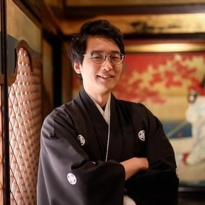 伝統的な黒い紋付袴を着用し、眼鏡をかけた男性が笑顔で立っているポートレート。背景には和風の装飾が見え、日本の文化や伝統を感じさせる一枚です。