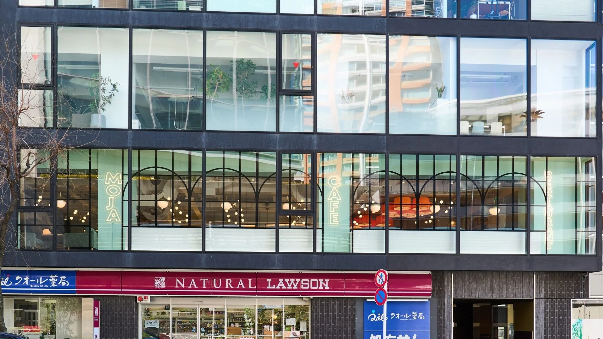 複数の店舗が入居する商業ビルの外観です。上層階には「MOJA CAFE RESTAURANT」のネオンサインが見え、下層階には「NATURAL LAWSON」と「るクオール薬局」が営業しています。現代的な都市の風景を捉えています。