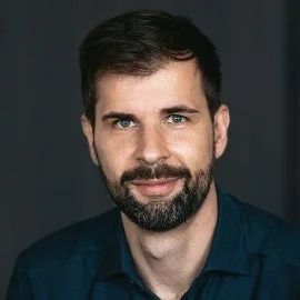 暗い背景を背に、ひげを生やした男性がカメラに向かって微笑んでいるポートレート写真です。青いシャツを着用しており、親しみやすくプロフェッショナルな印象を与えます。