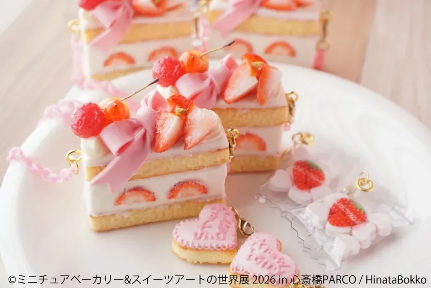 可愛らしいミニチュアのいちごショートケーキのチャームが複数並んでいます。ピンクのリボンやいちごで飾られ、ハート型のクッキーや包まれたいちごのお菓子も添えられています。スイーツアート作品の展示のようです。