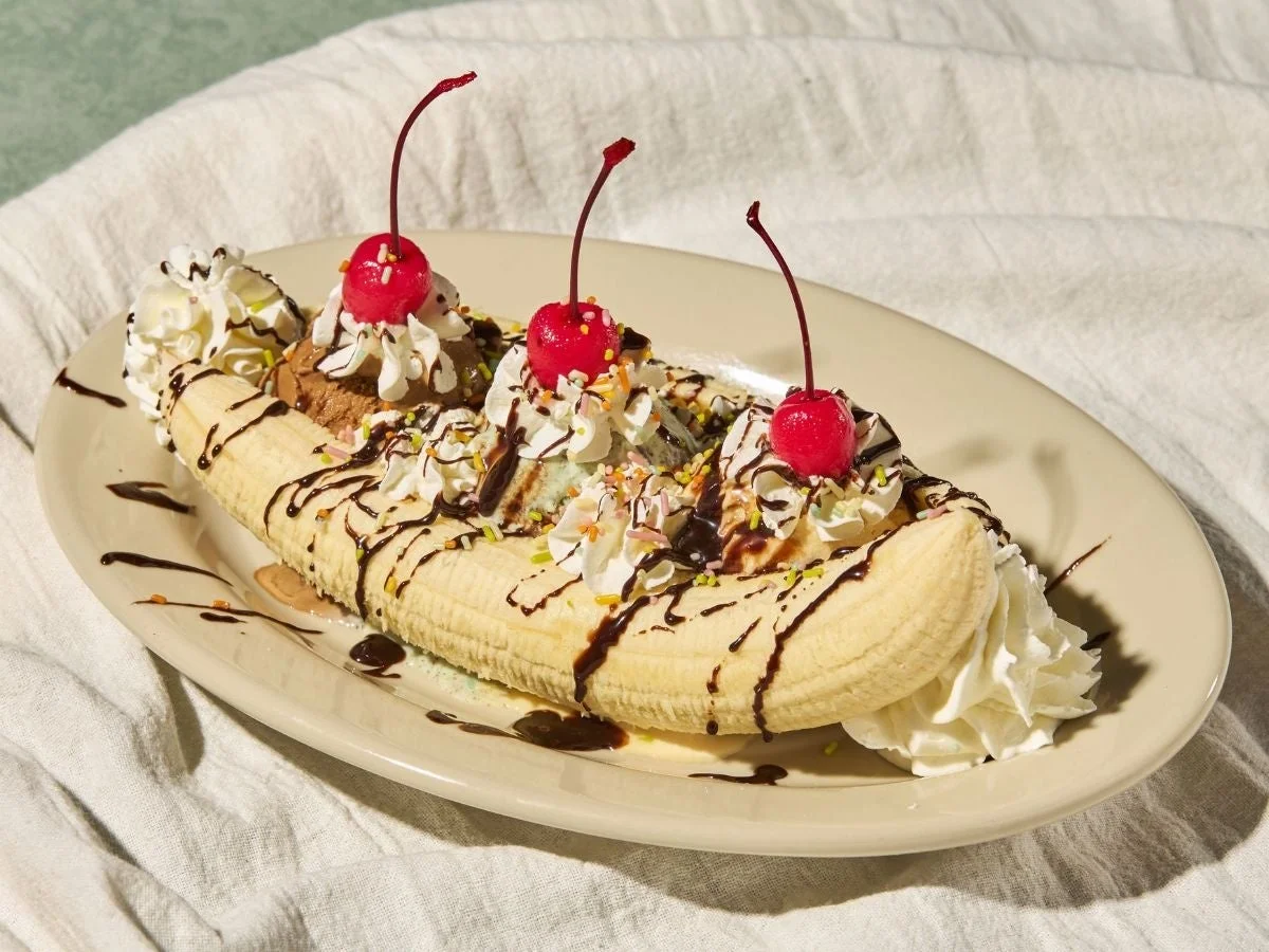 バナナを半分にカットし、数種類のアイスクリーム、たっぷりのホイップクリーム、チョコレートソース、カラースプレー、そして鮮やかな赤いチェリーがトッピングされた豪華なバナナスプリットです。