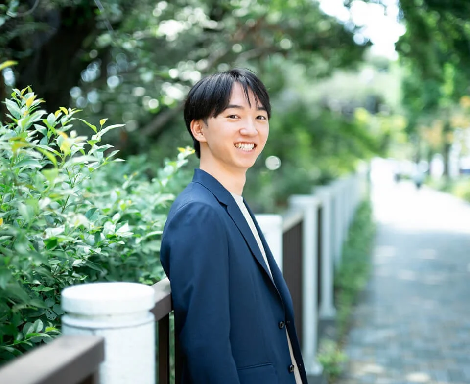 紺色のジャケットを着た若い男性が、緑豊かな屋外でカメラに向かって明るい笑顔を見せているポートレート写真です。爽やかな印象を与えます。