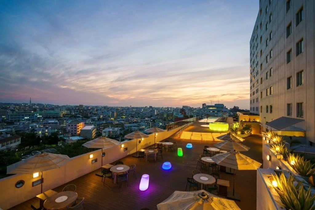 夕暮れ時の屋上テラス。テーブルや椅子が並び、遠くには街の明かりが見える